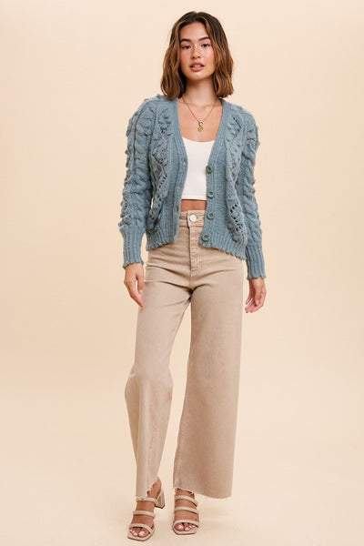 This feminine hand knit sage green cardigan is so sweet. Featuring a cable knit with pom poms and scalloped bottom.  Pairs perfectly with a white tee and denim for a instantly chic look.   Product Details:  50% Acrylic, 28% Recycled Polyester, 22% Polyester Cardigan with button front closure Shorter length
